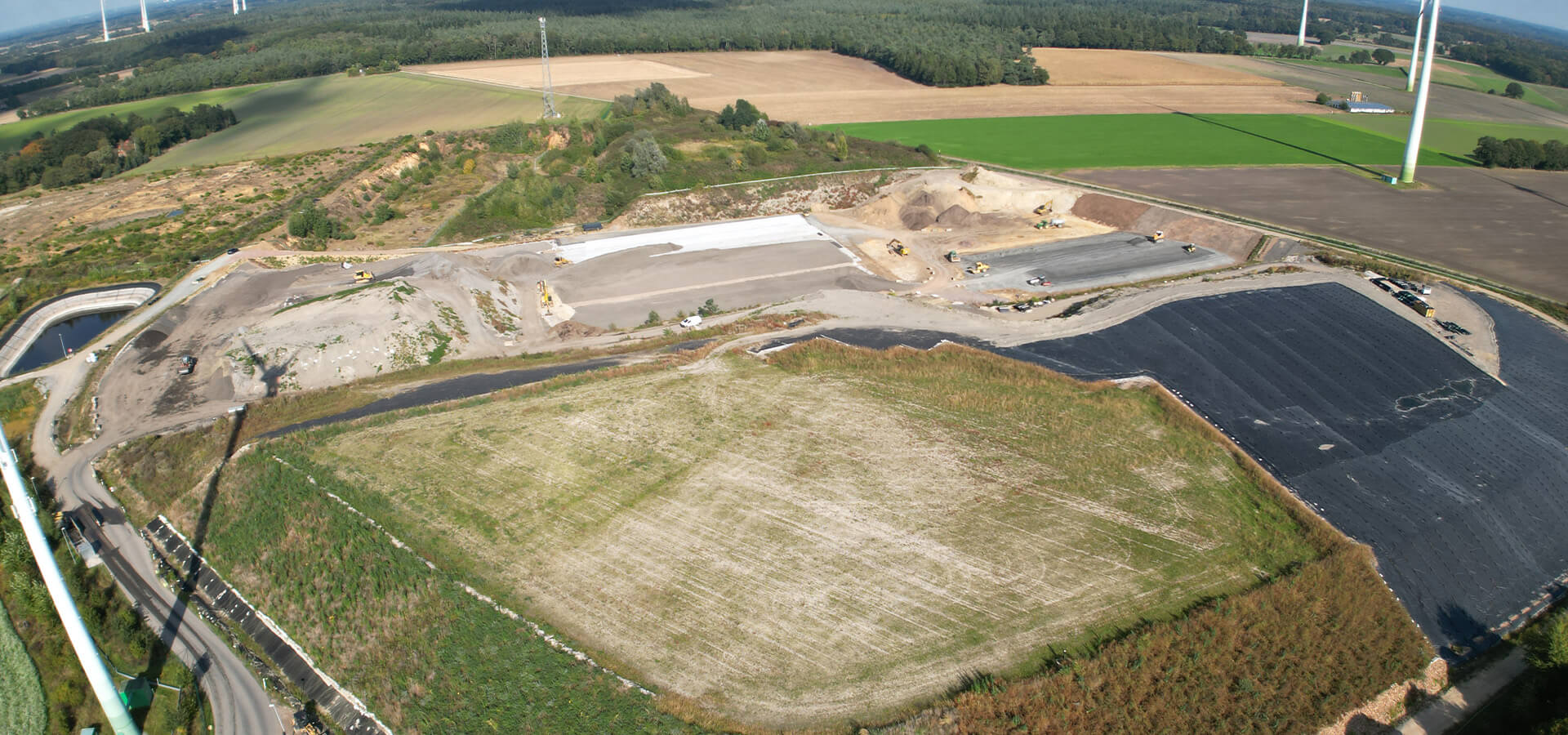Drohnenaufnahme mit neutraler Stimmung einer Baustelle mit grauen Erdflächen und schwarzen Folienfeldern zwischen Feldern und Windrädern