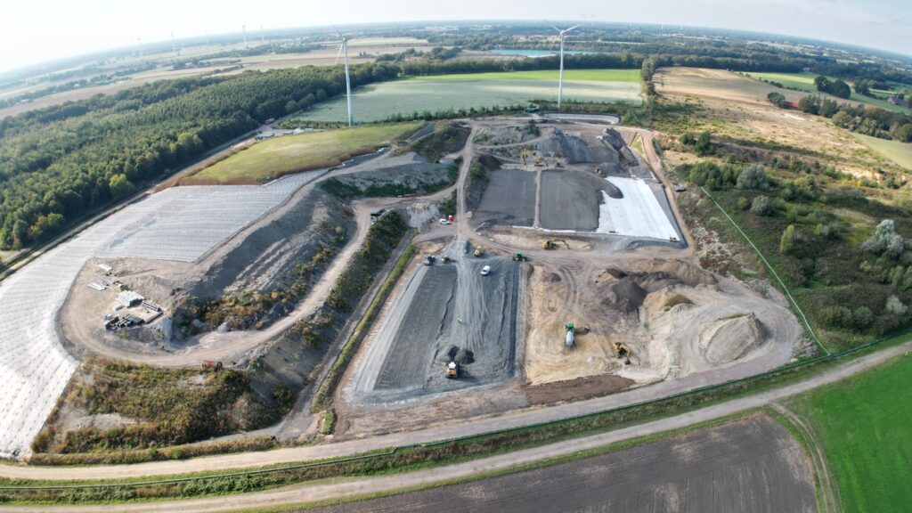 Luftbild einer Baustelle mit grauen Flächen und Windrädern in Grünland