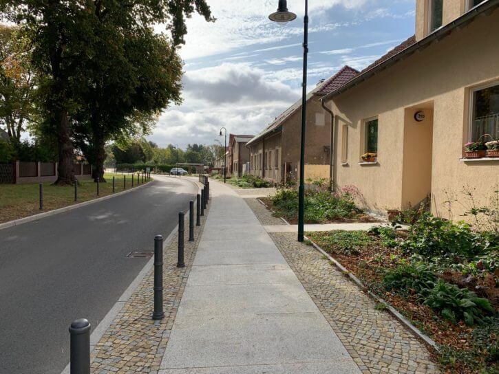 Leere Straße mit Gehweg, grauen Pollern, beige Häusern und Bäumen unter Wolkenhimmel