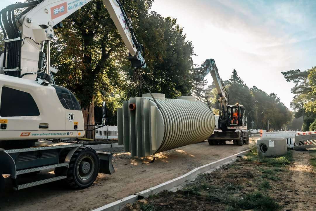 Zwei Bagger heben grauen Tank, Baustelle mit Bäumen, staubig, aktiv