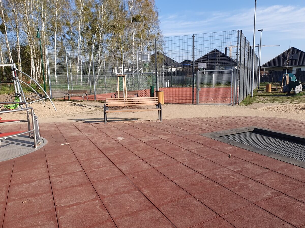 Leerer Spielplatz mit rotem Boden, Bank und eingezäuntem Sportplatz im Hintergrund