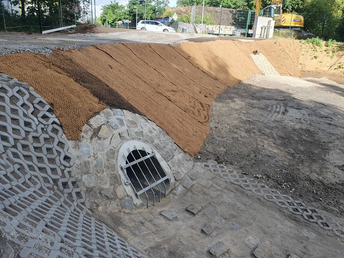 Brauner Erdwall mit Steinauslass und Metallgitter neben grauer Betonrinne